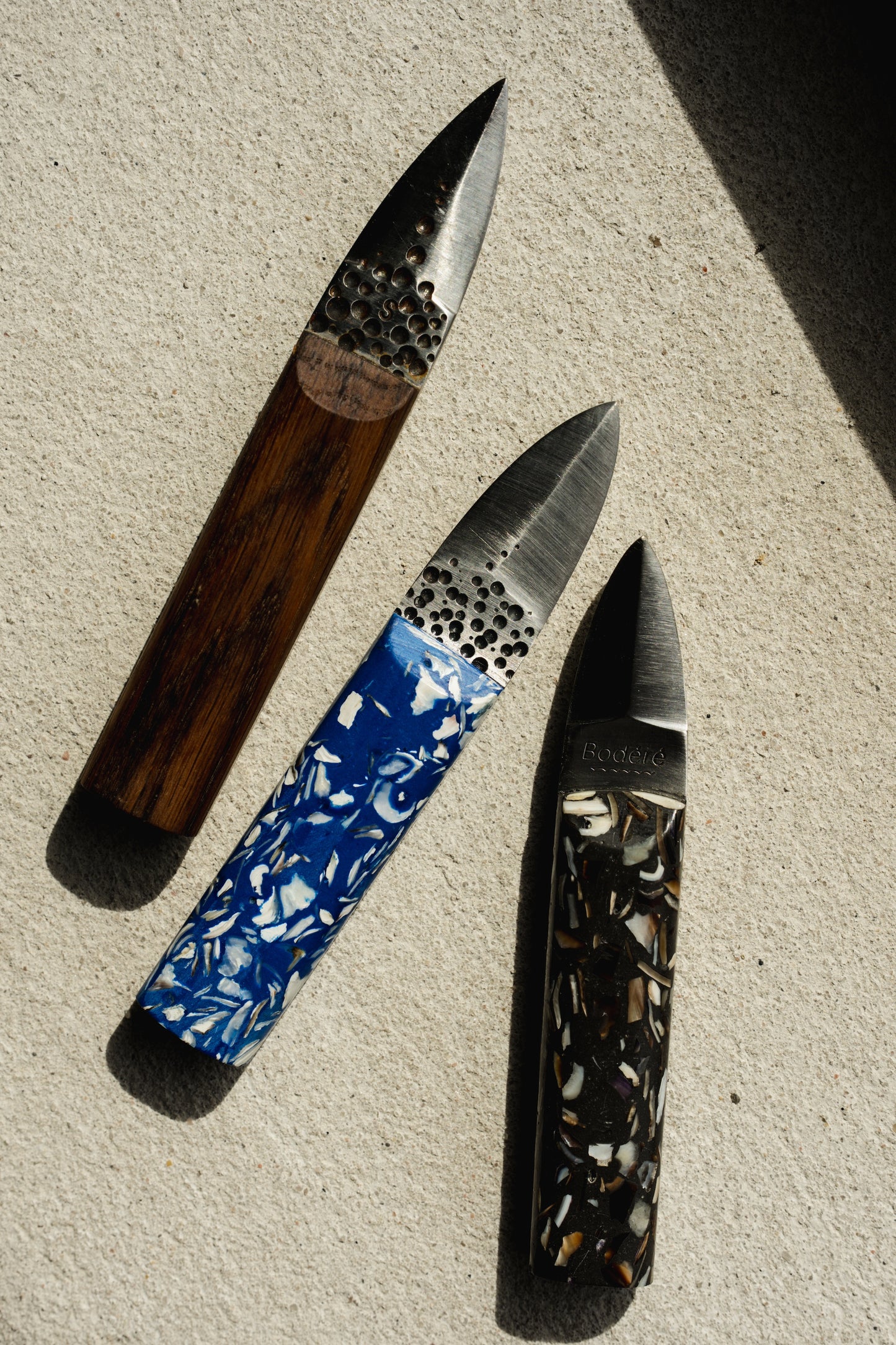 Couteaux à huîtres artisanaux bretons par Bodéré, avec manche en bois de chêne breton, en coquilles d'huîtres bleus et coquilles de moules noies sur un fond en béton naturel.