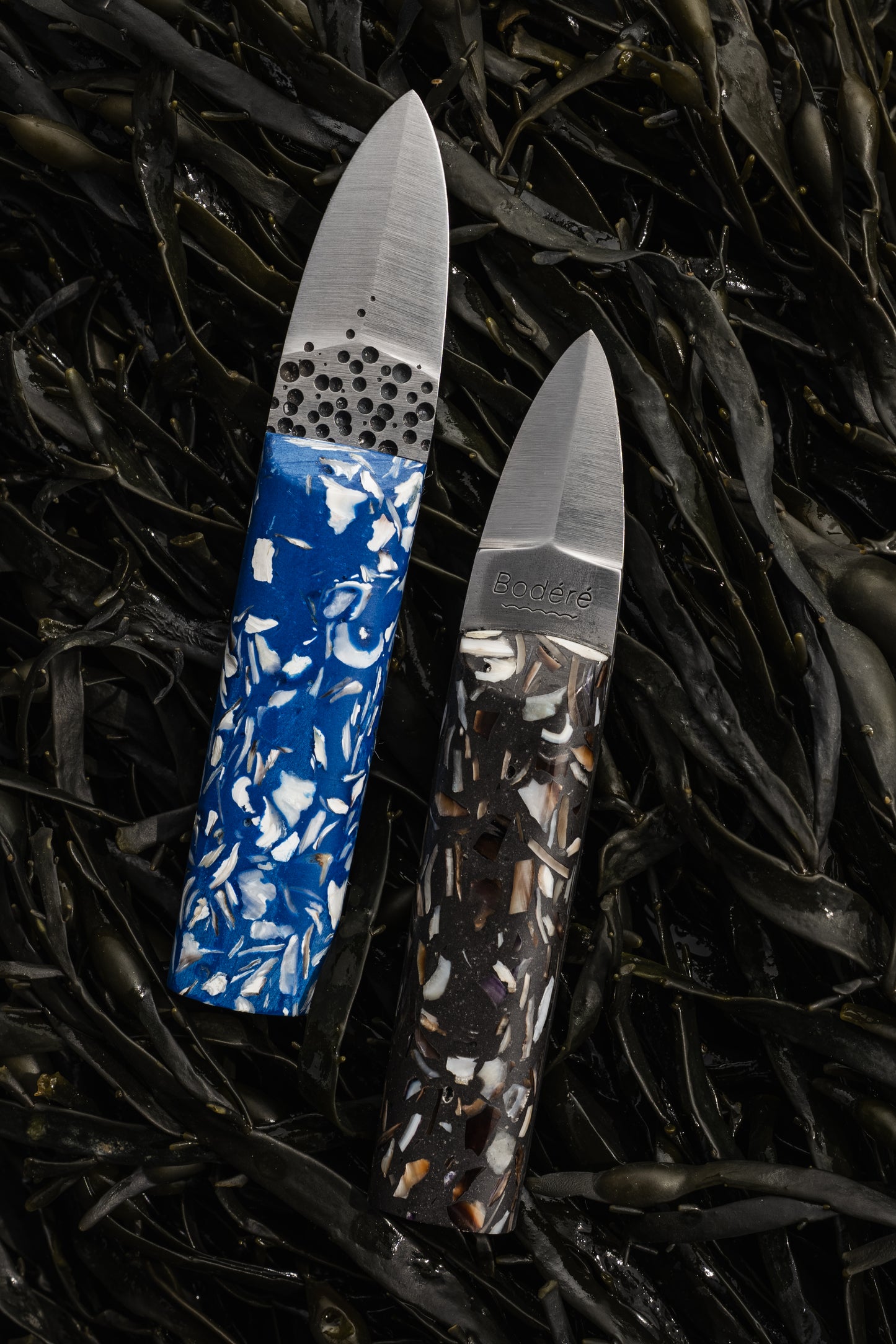 couteaux à huîtres artisanaux avec manches ecoresponsables bleus et noirs en coquillages recyclés sur un fond d'algues