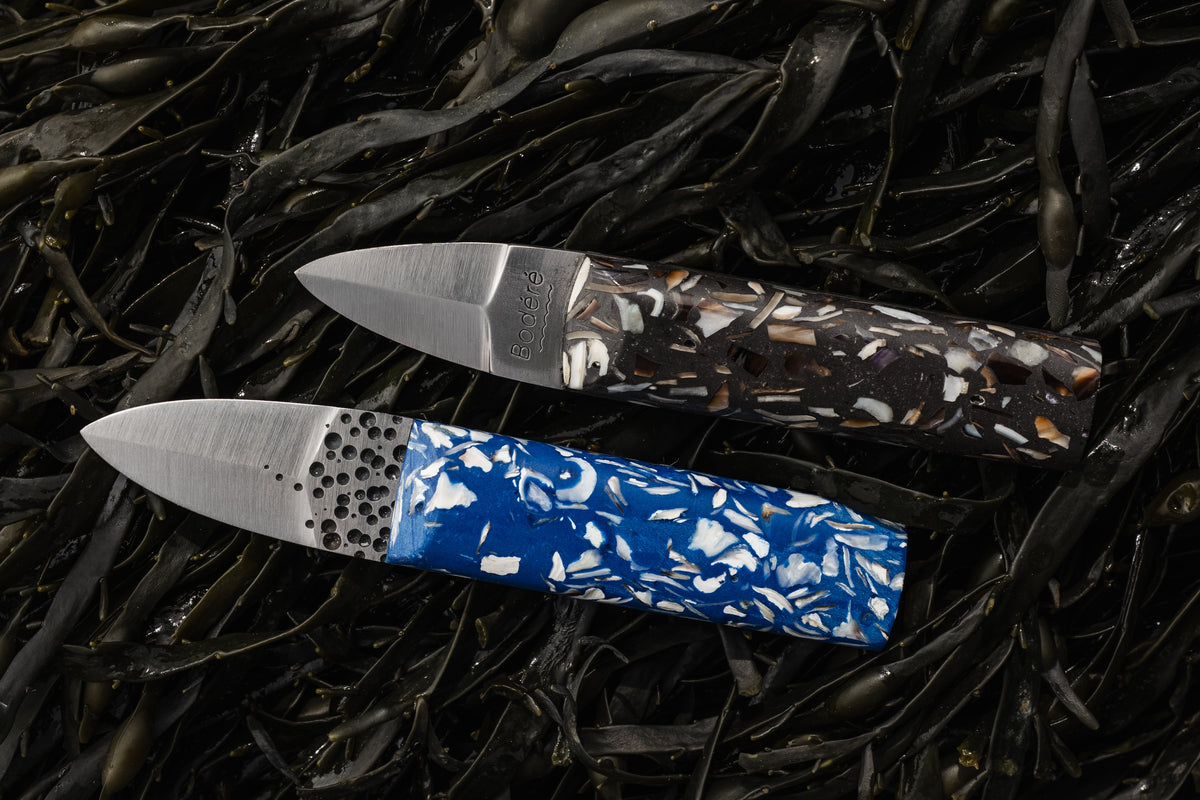 couteaux à huîtres artisanaux avec manches ecoresponsables bleus et noirs en coquillages recyclés sur un fond d'algues
